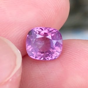 May include: A cushion-cut pink sapphire gemstone with a light purple hue.