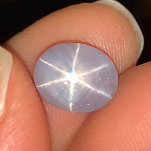 May include: A light blue oval gemstone with a six-pointed star pattern. The star is white and radiates from the center of the stone. The gemstone is held between two fingers.