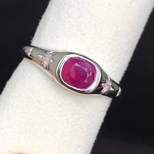 Puede incluir: Un anillo de tono plateado con una piedra preciosa central de color rojo rosado intenso, talla cojín, engastada en un bisel liso. La banda está decorada con dos motivos de estrellas grabados, cada uno con una pequeña piedra clara brillante.