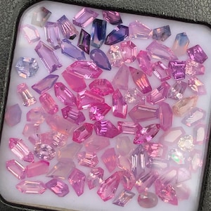 May include: A selection of cut gemstones in pink, purple, and clear hues. The gemstones are in diverse shapes, such as rectangular, oval, and teardrop, and are arranged in a white tray. The gems vary in size and colour intensity.