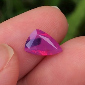 May include: A triangular cut pink opal gemstone with a purple and black inclusion. The gemstone is set in a clear setting and is held in a hand.