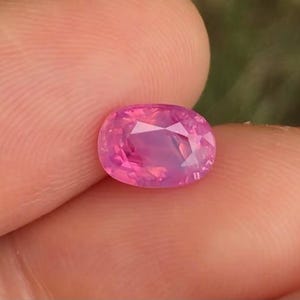 May include: A pink oval-shaped gemstone with a faceted cut. The gemstone is set against a light pink background.
