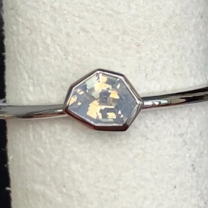 Puede incluir: Un anillo plateado con una piedra preciosa iridiscente hexagonal. La fina banda pulida sostiene una piedra con tonos azul p&aacute;lido, amarillo y dorado. El anillo se muestra sobre una superficie blanca.