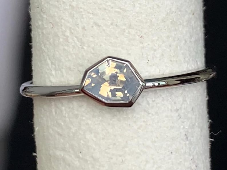 Puede incluir: Un anillo plateado con una piedra preciosa iridiscente hexagonal. La fina banda pulida sostiene una piedra con tonos azul p&aacute;lido, amarillo y dorado. El anillo se muestra sobre una superficie blanca.