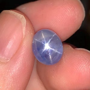 May include: A polished, oval blue star sapphire gemstone. The gem has a six-rayed star effect, with a bright, white star pattern radiating from the centre. The sapphire is held between fingers, showcasing its colour and clarity.