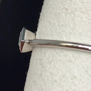 Puede incluir: Primer plano de un anillo de plata con una gema cuadrada de color azul claro. El anillo tiene una banda delgada y pulida, y la gema est&aacute; engastada en un engaste de cuatro puntas. El anillo se muestra sobre un fondo blanco.