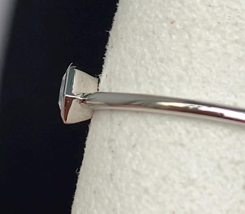 Puede incluir: Primer plano de un anillo de plata con una gema cuadrada de color azul claro. El anillo tiene una banda delgada y pulida, y la gema est&aacute; engastada en un engaste de cuatro puntas. El anillo se muestra sobre un fondo blanco.