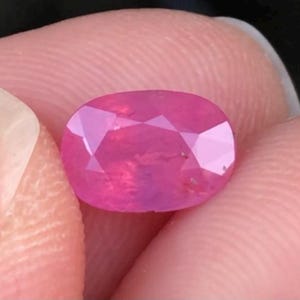 May include: A faceted, oval-cut ruby gemstone. The gem is a vibrant pinkish-red colour, with multiple facets that reflect light. The ruby is held between two fingers, showcasing its polished surface and internal clarity.