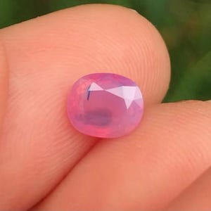May include: A pink oval gemstone with a faceted cut. The gemstone is held in a hand.