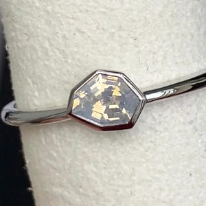 Puede incluir: Un anillo plateado con una gema hexagonal de color claro. La banda delgada est&aacute; pulida y la gema est&aacute; engastada en un bisel. El anillo se muestra sobre un fondo blanco.