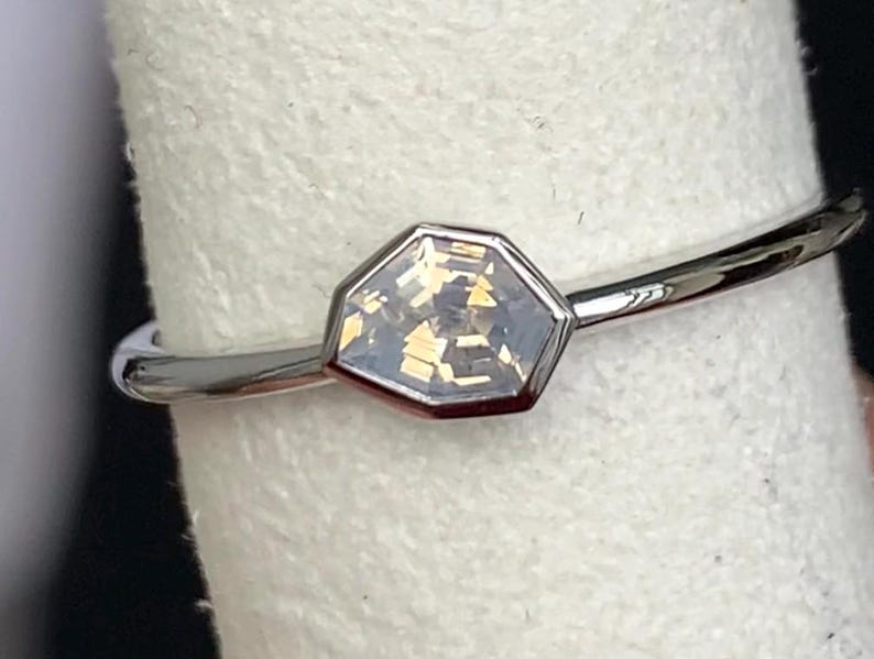 Puede incluir: Un anillo plateado con una gema hexagonal de color claro. La banda delgada est&aacute; pulida y la gema est&aacute; engastada en un bisel. El anillo se muestra sobre un fondo blanco.