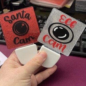 May include: Two decorative night lights with glittery square faces. One is red with "Santa Cam" text and a camera graphic, the other is silver with "Elf Cam" text and a camera graphic. Both have white bases.