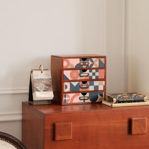May include: A wooden desk organizer with four drawers, each adorned with a unique geometric pattern in shades of pink, blue, and beige. The organizer sits atop a dark wooden dresser, with a calendar and a book nearby.