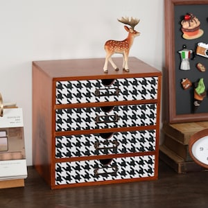 May include: A small wooden storage chest with four drawers. The drawer fronts feature a black and white houndstooth pattern. A small deer figurine with antlers sits on top. The chest is made of dark brown wood.