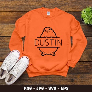 Peut inclure: Sweat-shirt orange vif avec un motif noir d'un hamster et le nom "Dustin". Le design est centré sur le devant du col rond à manches longues. Des baskets blanches sont placées en dessous.