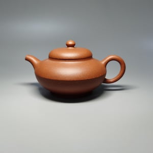 Może przedstawiać: Mały, brązowy ceramiczny imbryk z pokrywką i zakrzywionym uchwytem. Imbryk ma dzióbek do nalewania i zaokrąglony korpus. Kolor jest ciepły, ziemisty brąz, a powierzchnia wydaje się mieć lekko teksturowane wykończenie. Przeznaczony do parzenia herbaty.