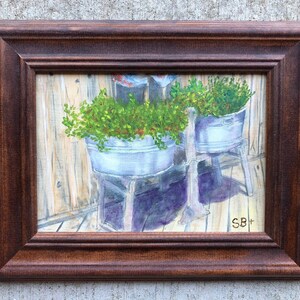 May include: An original painting in a brown wooden frame. The artwork depicts two galvanized metal buckets filled with green plants. The buckets sit on wooden stands against a weathered wooden background. The painting is signed in the lower right corner.