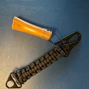 May include: A handmade lanyard with a black metal clip and a woven paracord strap in shades of green and black. The lanyard is attached to a wooden object encased in a clear plastic tube.