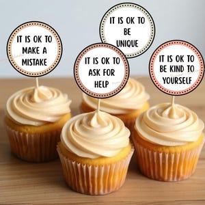 Könnte beinhalten: Vier Cupcakes mit hellbraunem Zuckerguss und dekorativen Toppern. Jeder Topper hat ein kreisförmiges Design mit einer positiven Aussage: „It is ok to make a mistake“, „It is ok to be unique“, „It is ok to ask for help“ und „It is ok to be kind to yourself“.