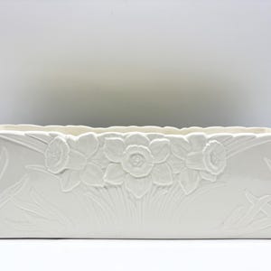 Può includere: Una fioriera rettangolare in ceramica color bianco sporco caratterizzata da un design in rilievo con tre fiori di narciso centrati sul pannello frontale, circondati da motivi a foglia in rilievo che si estendono lungo il contenitore.