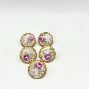 Puede incluir: Un conjunto de cinco botones circulares con un centro blanco y motivos florales de rosas rosas y blancas pintadas, rodeados por un borde dorado metálico decorativo con una textura de filigrana en relieve en el borde exterior.