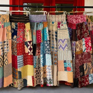 Puede incluir: Una fila de seis faldas largas colgadas en un perchero de metal negro. Cada falda presenta un diseño de patchwork único con varios patrones coloridos, incluyendo estampados florales, geométricos y de animales. Las faldas tienen cinturas elásticas.