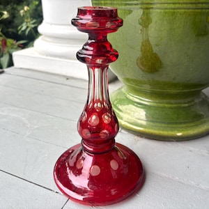 May include: A ruby red glass candlestick holder featuring a clear glass stem with vertical fluting and a bulbous base decorated with circular clear glass polka dot accents. The candlestick has a wide, circular foot and a flared top rim.