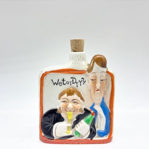 May include: A ceramic bottle with a cork stopper, featuring a rectangular design with a white background and orange trim. The bottle has two illustrated faces and the text "Wet or Dry?" on the front. One figure is smiling and holding a glass, while the other looks sad.