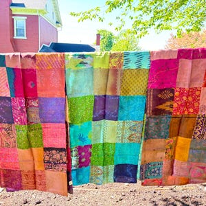 Op de afbeelding: Een kleurrijk patchwork textiel, waarschijnlijk een quilt of sprei, hangt buiten. De stof bestaat uit verschillende rechthoekige lappen in roze, oranje, groen, blauw en paars. Het textiel wordt gepresenteerd tegen een achtergrond van groen gras en een rood bakstenen gebouw.