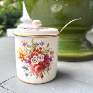 May include: A white porcelain jam jar with a matching lid and a small metal spoon. The jar features a colorful floral pattern with red, pink, yellow, and blue flowers and green leaves. The rim of the jar and the lid are accented with gold trim.