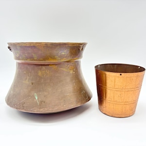 May include: Two antique copper vessels. One is a large, flared pot with a wide rim and a patinaed finish. The other is a smaller, cylindrical bucket with horizontal bands. Both have a warm, reddish-brown color.