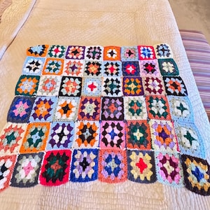May include: A square crochet blanket composed of numerous multi-colored granny squares. Each individual square features a central color surrounded by contrasting yarn borders in various shades including red, orange, yellow, green, blue, purple, and white.