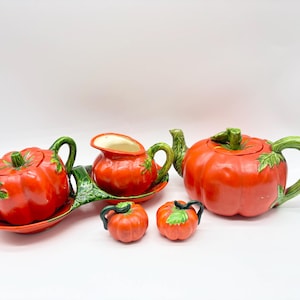 May include: A ceramic tea set with a tomato theme. The set includes a teapot, creamer, sugar bowl, and two small tomato-shaped containers. The teapot and creamer have green handles and leaf accents. The set is primarily red and green.
