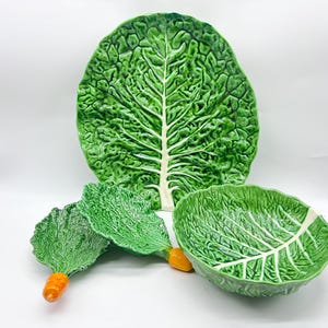 May include: A collection of ceramic serving dishes shaped like cabbage leaves. The set comprises a large platter, a bowl, and two leaf-shaped dishes, all in green with white veining. The stems of the smaller dishes are orange.