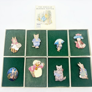 May include: A set of eight enamel pins featuring characters from "The World of Beatrix Potter," presented in individual green boxes. The pins depict various animals in colourful outfits, with a book of the same title displayed above.