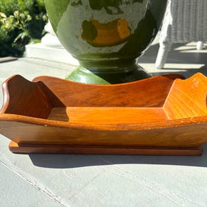 May include: A rectangular wooden tray with a warm brown finish, featuring curved, elevated sides and a flat base. The tray has a smooth, polished surface with visible wood grain patterns and a slightly flared rim design along the edges.