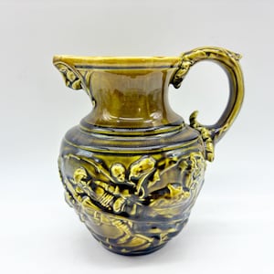 May include: A glazed ceramic pitcher in a deep olive green color featuring a relief design of figures in motion around the body. The vessel has a wide spout, a curved handle with ornate detailing, and a smooth, glossy finish throughout.