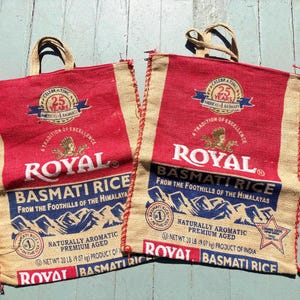 Könnte beinhalten: Zwei Tragetaschen aus Jute mit roten und beigen Paneelen. Die Taschen zeigen den Text "ROYAL BASMATI RICE" und ein "25 YEARS" Emblem. Die Taschen bestehen aus einem rauen, gewebten Material und haben Seilgriffe.