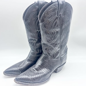 Puede incluir: Un par de botas vaqueras de cuero negro con punta afilada, tacón de bloque y patrones de costura tonales intrincados en la caña alta. El empeine y la puntera muestran un acabado texturizado inspirado en reptiles con un brillo lustroso.