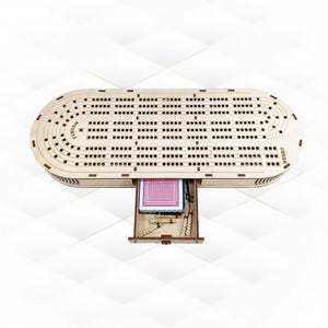 Può includere: Un tabellone da cribbage in legno con un cassetto per riporre le carte. Il tabellone ha un design tradizionale con spazi numerati e un traguardo. Il cassetto è aperto e contiene un mazzo di carte.