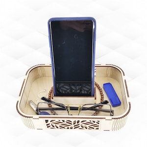 May include: A wooden organiser with a carved leaf design. It has a slot for a phone, a small compartment for a blue case, and a space for glasses and jewellery.