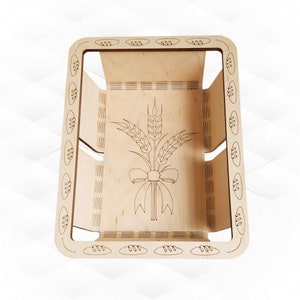 Op de afbeelding: Een houten broodmand met een decoratief ontwerp van tarwehalmen en een strik. De mand is lichtbruin van kleur en heeft een rechthoekige vorm.
