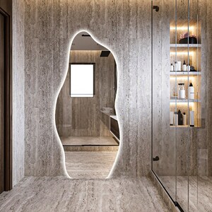 Op de afbeelding: Een badkamer met een grote, onregelmatig gevormde spiegel met een licht rond de rand. De spiegel reflecteert een douche met een glazen deur en een raam. De muren zijn gemaakt van lichtbruine steen.