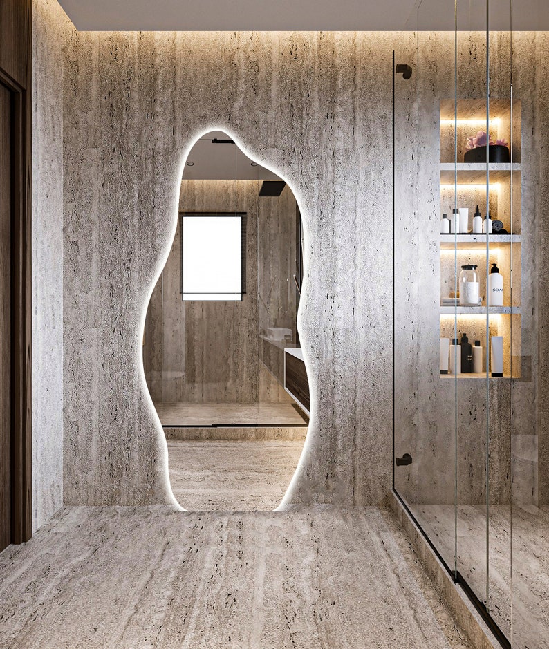 Op de afbeelding: Een badkamer met een grote, onregelmatig gevormde spiegel met een licht rond de rand. De spiegel reflecteert een douche met een glazen deur en een raam. De muren zijn gemaakt van lichtbruine steen.