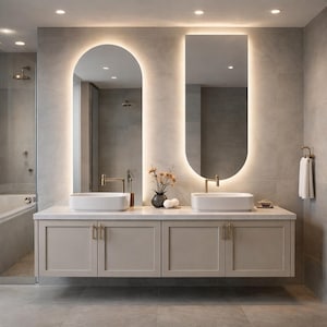 May include: Modern bathroom design featuring a floating vanity with two white rectangular sinks and gold fixtures. Two illuminated mirrors, one arched and one oval, are mounted above the vanity. The color scheme is neutral with gray tiles and a light beige vanity.