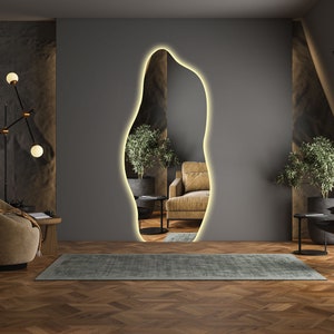 Op de afbeelding: Een modern woonkamerinterieur met een grote, organisch gevormde spiegel met een warm witte lichtcontour. De spiegel bevindt zich op een grijze muur, en de kamer heeft een houten vloer met een visgraatpatroon. Op de voorgrond staat een bruine fauteuil, op de achtergrond een kleine plant in een pot.