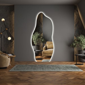 Op de afbeelding: Een moderne woonkamer met een grote, organisch gevormde spiegel met een witte omtrek. De spiegel reflecteert een bruine fauteuil en een plant in een pot. De kamer heeft een donkere houten vloer, een bruine fauteuil en een zwarte open haard.