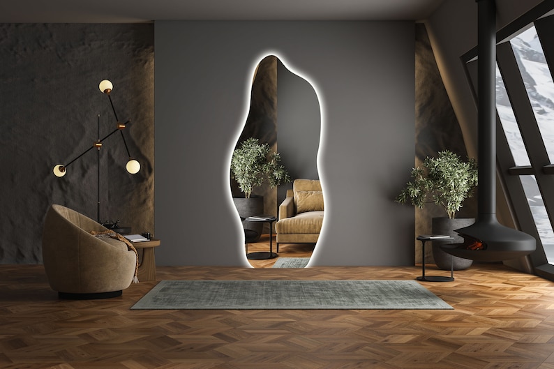 Op de afbeelding: Een moderne woonkamer met een grote, organisch gevormde spiegel met een witte omtrek. De spiegel reflecteert een bruine fauteuil en een plant in een pot. De kamer heeft een donkere houten vloer, een bruine fauteuil en een zwarte open haard.