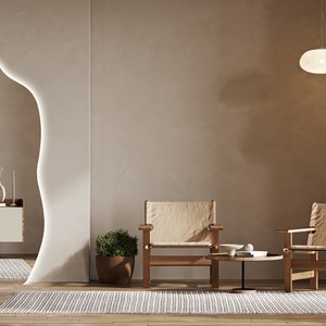 Op de afbeelding: Twee beige stoelen met houten frames en een kleine ronde tafel met een witte keramische kom en een boek erop. De stoelen en de tafel staan voor een beige muur met een grote, organisch gevormde spiegel met een witte omtrek. Twee witte hanglampen hangen aan het plafond.