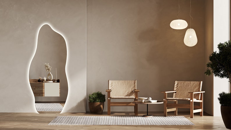 Op de afbeelding: Twee beige stoelen met houten frames en een kleine ronde tafel met een witte keramische kom en een boek erop. De stoelen en de tafel staan voor een beige muur met een grote, organisch gevormde spiegel met een witte omtrek. Twee witte hanglampen hangen aan het plafond.